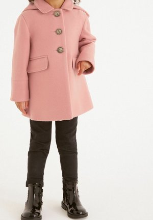 Niña vestida con un abrigo rosa abotonado con bolsillos de solapa, pantalones negros y botines negros brillantes, de pie sobre un fondo blanco.