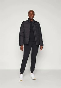 Melnā pūkainā jaka ar rāvējslēdzēju priekšā un augstu apkakli, valkāta virs melna krekla, apvienota ar melnām šaurām biksēm un baltām sporta apaviem.