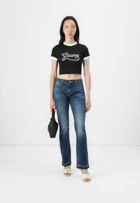 Svart kortermet crop top med hvite detaljer som har "Guess"-logo, parret med blå vide jeans og beige slip-on sandaler. Svart håndveske.