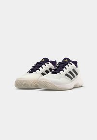 Weiße Sportschuhe mit einer strukturierten Oberfläche, lila Akzenten, dunklen Schnürsenkeln und drei schwarzen Streifen. Die Sohle hat ein Fischgradmuster.