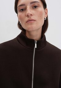 Veste zippée marron avec un col haut. Tissu doux, design minimaliste, détail de fermeture éclair en argent, sans logos ni motifs visibles.