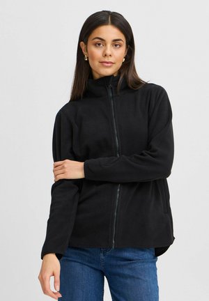 Giacca nera in pile con zip, colletto alto, maniche lunghe e tasche laterali, abbinata a jeans blu. Tessuto morbido, fit casual.