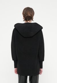 Casaco de fleecy preto com capuz, com um corte descontraído, mangas largas e uma superfície texturizada. A bainha cai abaixo dos quadris.