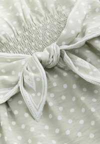 Hellgrüner Stoff mit weißen Tupfen, ausgestattet mit gesammeltem Gummizug an der Taille und einer gebundenen Schleife zur Anpassung. Glatte Textur.