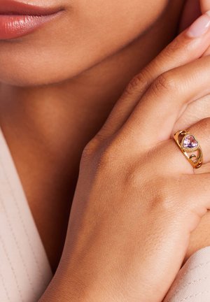 Gouden ring met een hartvormige roze edelsteen, voorzien van een draaiend ontwerp. Hand met een gladde huid verbergt gedeeltelijk de ring.