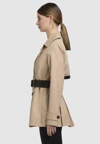 Beige trenchcoat met een getailleerd ontwerp, zwarte riem, brede kraag en lange mouwen. Bevat knoopaccenten en een geplooide achterkant.