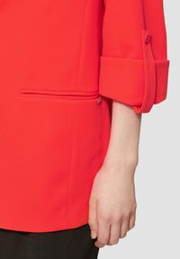 Blazer rouge avec une texture lisse, présentant des manches retroussées et une petite poche latérale. Le détail des boutons sur la manche ajoute une touche fonctionnelle.