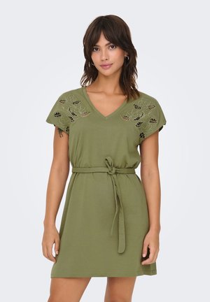 Femme portant une robe courte vert olive avec des manches découpées florales et une ceinture en tissu à la taille, debout devant un fond clair uni.