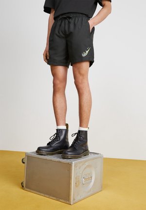 Persoon met zwarte korte broek en zwarte veterlaarzen staat op een zilveren metalen koffer op een gele vloer tegen een effen witte muur.