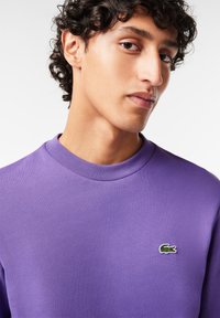 Lilla sweatshirt med rund halsudskæring, lavet af glat stof. Har et lille grønt og hvidt krokodillelogo på venstre bryst.