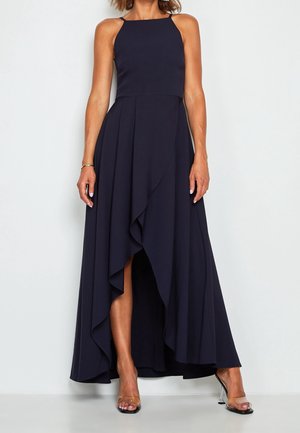 Woman wearing a sleeveless navy blue high-low dress with ruffled front hem and clear high-heeled sandals, standing against white background.