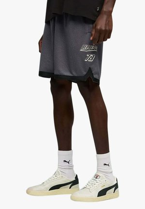 Persona che indossa pantaloncini grigi in mesh con la scritta "REPRESENT 73", calzini bianchi Puma e sneaker crema Puma con dettagli neri.