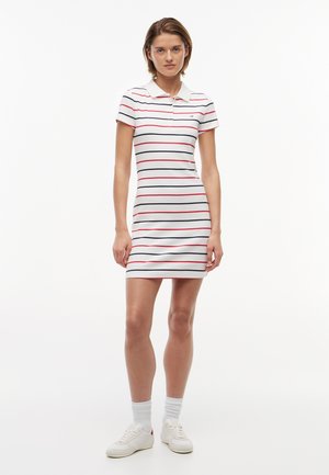Frau steht und trägt ein weißes Polokleid mit kurzen Ärmeln und roten sowie dunkelblauen horizontalen Streifen, weiße Socken und weiße Turnschuhe.
