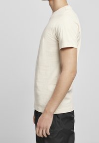 Beige bavlněné tričko s krátkým rukávem, kulatým výstřihem a volným střihem, nošené s černými šortkami. Hladká textura a minimalistický design.