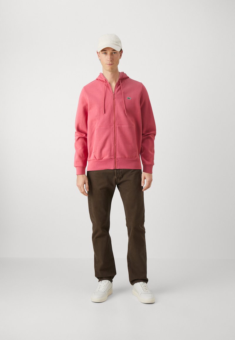Haut zippé rose avec une poche kangourou, porté sur un pantalon marron et des baskets blanches. Le hoodie présente un petit logo sur la poitrine gauche.