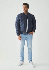 Veste matelassée navy avec fermeture éclair à l'avant et poches latérales, associée à un jean bleu clair et des baskets blanches. Tissu lisse et coupe moderne.