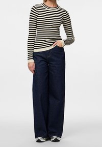 Femme portant un haut à manches longues rayé noir et blanc, jean large sombre, baskets blanches, debout avec une main dans la poche.