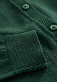 Grüne Strickjacke aus weichem Strickmaterial, mit gerippten Bündchen und passenden grünen Knöpfen mit Kreuzdesign.