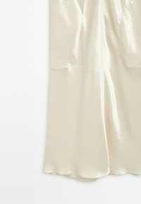 Cream-colored silky fabric with a subtle floral texture and a central seam, forming a flowing garment hem.