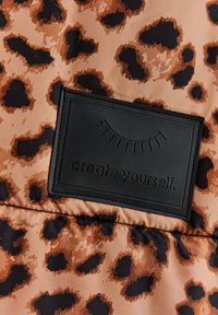 Tkanina s leopardím vzorom a čiernou nášivkou s grafickým zobrazením rias a textom "vytvor si seba."