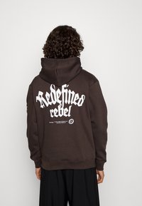 Mørkebrun hættetrøje med stort hvidt tekst "Redefined rebel" på ryggen, med ribbede manchetter og kant, samt en frontlomme med kenguru-åbning.