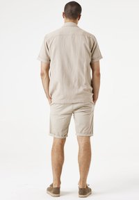 Beige shirt met korte mouwen met een textuurpatroon, gecombineerd met licht kaki shorts. Het model draagt bruine, suède-achtige schoenen.