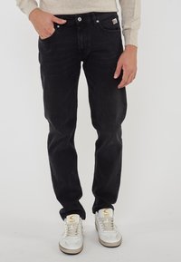 Uomo che indossa jeans neri con una piccola etichetta del marchio e sneakers bianche con dettagli beige, in piedi di fronte a uno sfondo semplice.