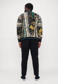 Veste bomber multicolore avec des motifs abstraits en noir, jaune, vert et blanc. Manches et ourlet côtelés, associée à un pantalon noir.