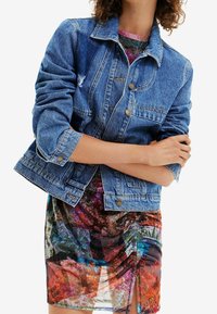 Denim jacket with a classic cut, featuring silver buttons and a light wash, layered over a colorful floral-printed dress.