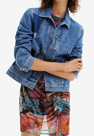 Veste en denim à coupe classique, dotée de boutons argentés et d'un délavage léger, superposée à une robe à imprimé floral coloré.