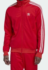 Red adidas zip-up tracksuit jacket with high collar, white three-stripe accents on sleeves, and ribbed cuffs. Soft fabric texture.
