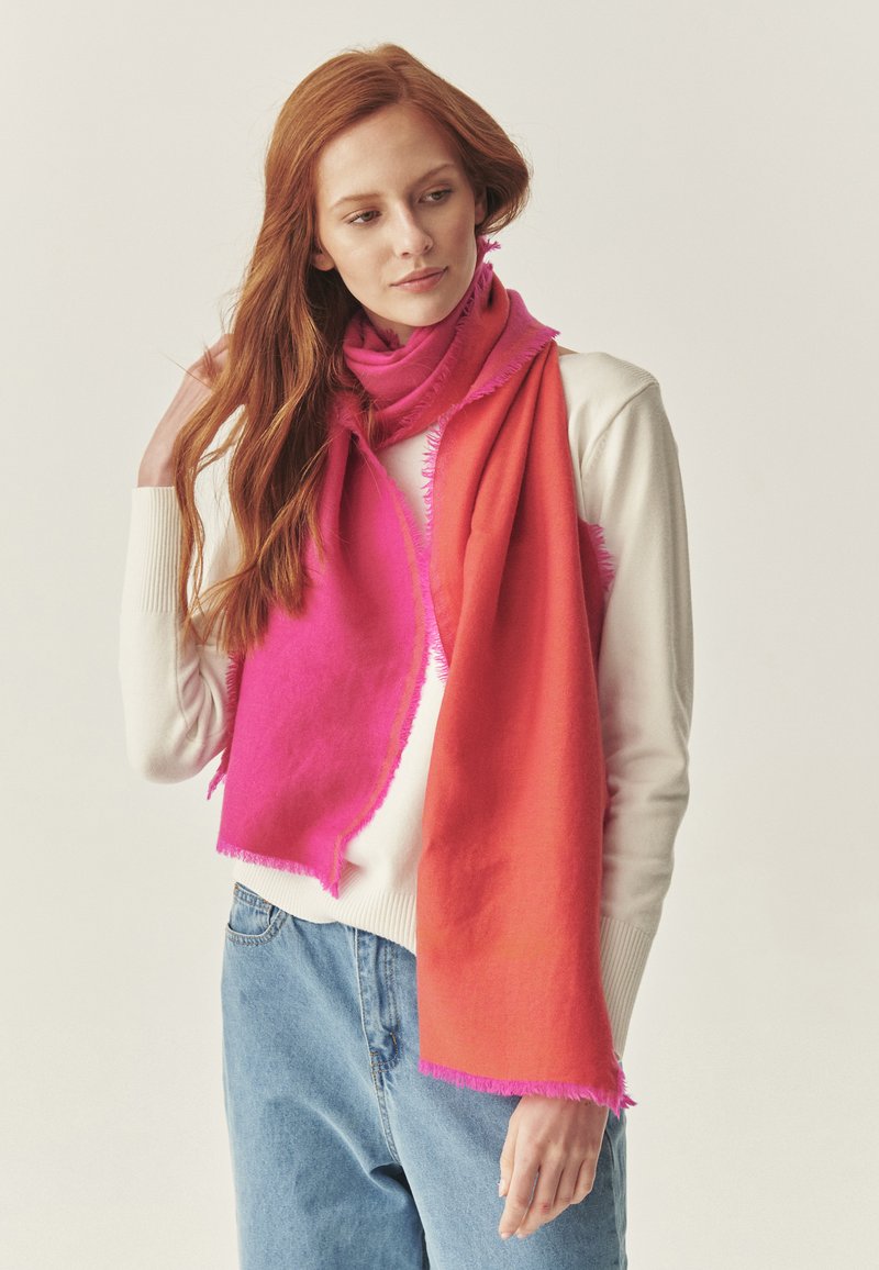 Sciarpa bicolore con tonalità vivaci di rosa e arancione, con una texture morbida e bordi sfilacciati. Indossata drappeggiata attorno al collo.