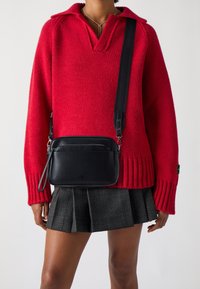 Borsa a tracolla nera in pelle con superficie texturizzata, tasche con zip, forma piatta e tracolla regolabile, indossata sopra un maglione rosso e una gonna a quadri.