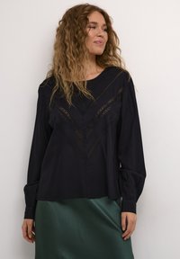 Blouse marine à manches longues et décolleté en V. Présente des détails en dentelle dans un motif en chevrons. Portée avec une jupe verte texturée.