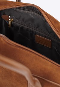 Binnenkant van een bruine suede handtas met zwarte voering, voorzien van een ritsvak en leren merketiket. Gladde textuur en ruim ontwerp.