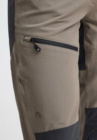 Khaki buitenbroek met een gestructureerde afwerking, voorzien van een zwarte patch aan de zijkant en een met rits afsluitbare pocket. Centraal logo in donkerbruin.