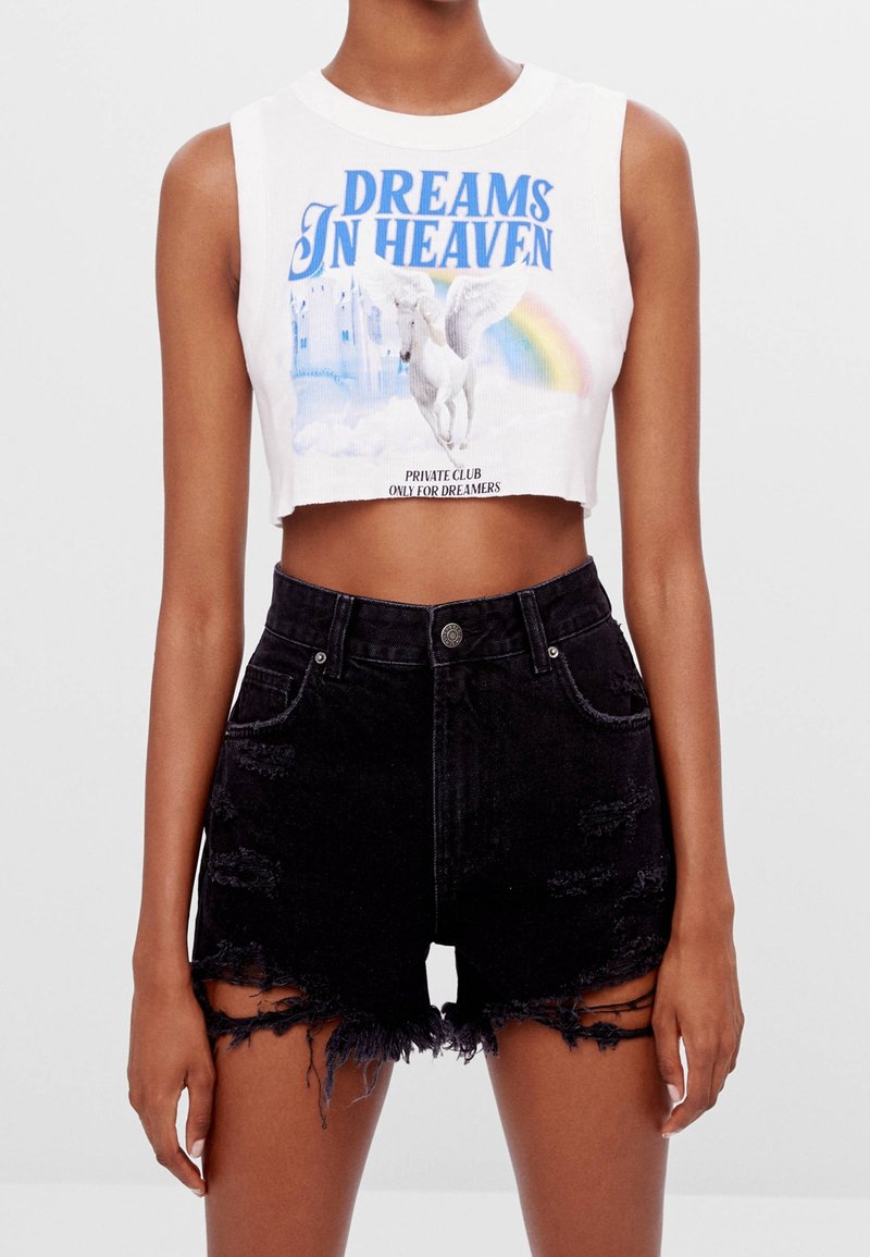 Crop top blanc avec un graphisme bleu "DREAMS IN HEAVEN" mettant en scène une licorne et un arc-en-ciel, associé à un short en denim noir avec des bords effilochés.
