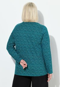 Långärmad topp i turkos med en subtil mönstrad textur. Rak skärning, solid bakvy. Parat med svarta byxor.