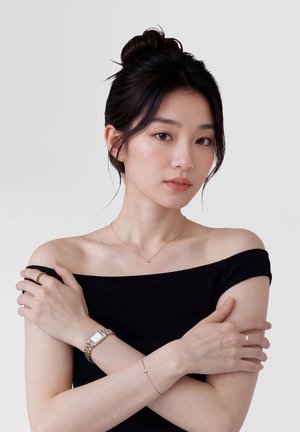 Tânără cu top negru pe umeri, părul prins într-un coc, poartă un ceas, un colier delicat, o brățară și inele, cu brațele încrucișate pe piept.