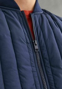 Giacca bomber imbottita blu navy con zip frontale, colletto a coste e fodera rossa a contrasto, caratterizzata da una superficie liscia e un delicato lucido.