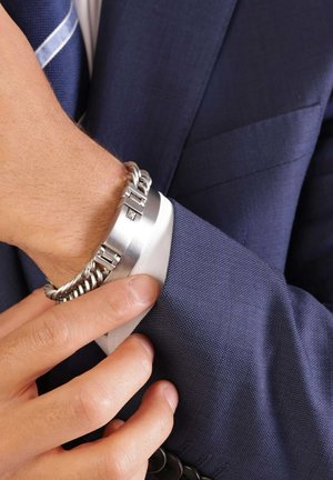 Silver metal bracelet with a polished finish, featuring a chain link design and a rectangular clasp. Worn over a dark blue suit sleeve.