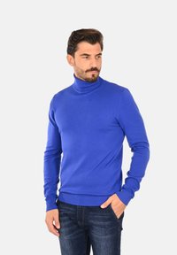 Maglione turtleneck blu realizzato in tessuto a maglia, con un design aderente e bordi a coste. Abbinato a jeans in denim scuro.