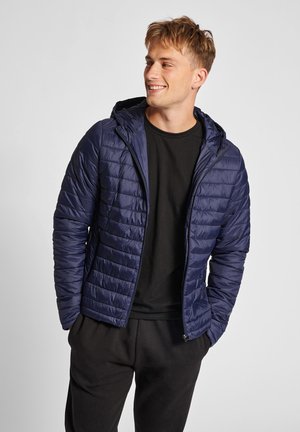 Marineblauwe gewatteerde jas met een ritssluiting en capuchon. Voorzien van een diagonaal doorgestikte patroon en elastische manchetten. Gecombineerd met een zwart t-shirt en donkere broek.