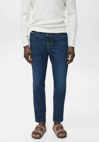 Dunkelblaue Denim-Jeans mit geradem Schnitt, ausgestattet mit fünf Taschen und kontrastierenden Nähten, kombiniert mit beigen Sandalen und einem gerippten weißen Pullover.