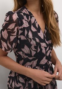 Musta mekko, jossa vaaleanpunaisia lehtikuvioita, läpikuultava kangas ja V-pääntie. Mekossa on lyhyet, pussihihat ja vyötäröosassa laskokset.