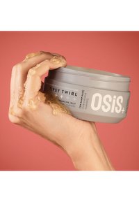 Grå behållare med hårgelé märkt "OSiS+ TIPSY TWIRL," hålls i en hand med gelé som droppar. Bakgrunden är en enfärgad korallfärg.