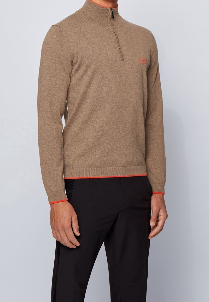 Man i en ljusbrun zip-up tröja med orange kant vid ärmar och nederkant, tillsammans med svarta byxor framför en neutral bakgrund.