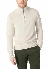 Mann in einem cremefarbenen Strickpullover mit Reißverschlusskragen und dunkelgrünen Hosen, mit einer Hand in der Tasche.