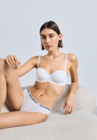 Bílá tvarovaná podprsenka s tenkými ramínky a sladěné kalhotky s černým páskem s logem, vystavené na strukturovaném světle zbarveném povrchu.