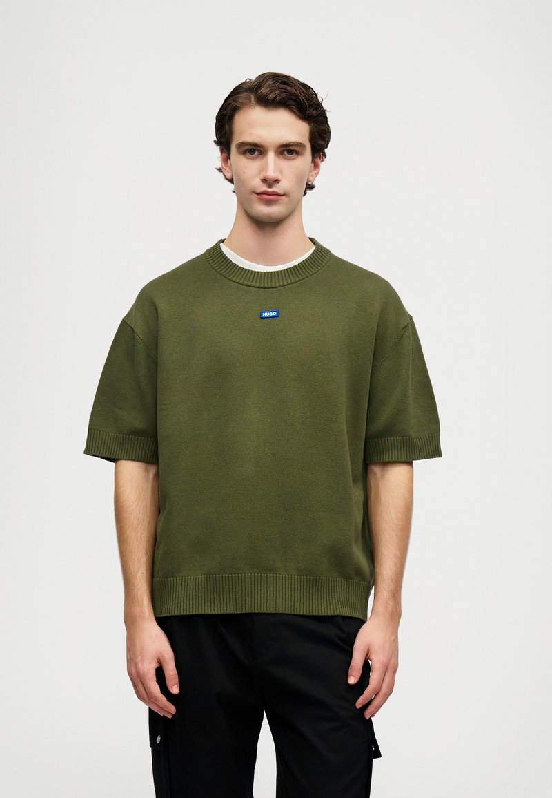 Ελαιόχρωμο κοντομάνικο πλεκτό πουλόβερ με ριμπ μανσέτες και στρίφωμα, διαθέτει έναν μικρό μπλε λογότυπο στο στήθος. Λείος υφασμάτινος χαρακτήρας και άνετη εφαρμογή.